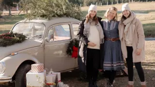 Drei Frauen posieren neben einem Oldtimer, der mit einem Weihnachtsbaum geschmückt ist, umgeben von verpackten Geschenken und Weihnachtsdekoration.