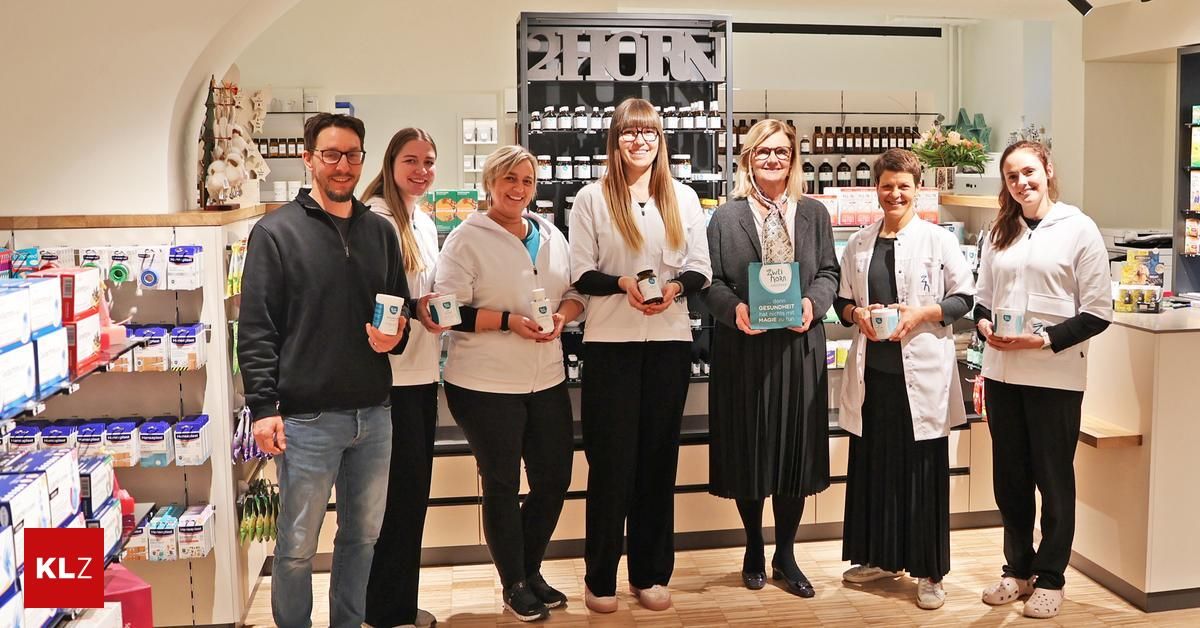 Five individuals, including two women in white coats, stand in a pharmacy. They hold cups and display a certificate, smiling at the camera. Shelves filled with bottles are in the background.