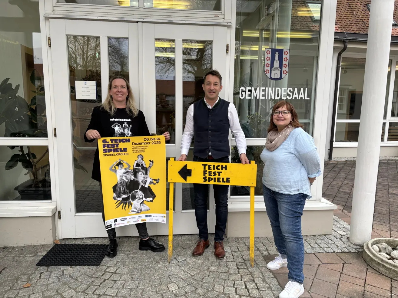 Drei Personen stehen vor einem Gebäude mit einem Schild, das 'Teich Fest Spiele' besagt. Die Frau auf der linken Seite hält ein gelbes Plakat. Der Mann in der Mitte trägt eine Weste und ein Lächeln. Die Frau auf der rechten Seite trägt eine Brille und einen Schal. Hinter ihnen befindet sich eine Glastür mit einem Logo.