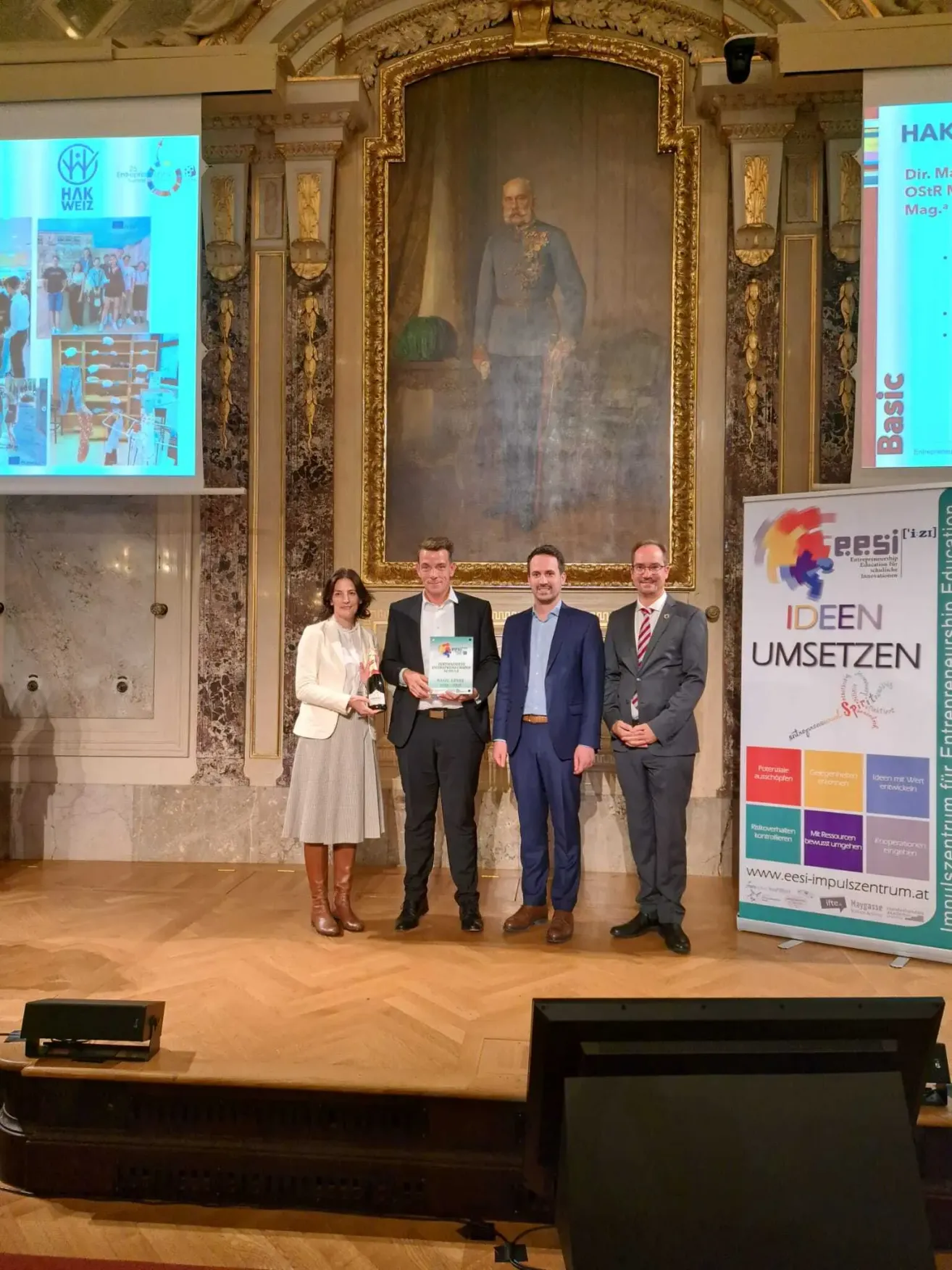 Vier Personen stehen auf einer Bühne und erhalten eine Auszeichnung. Eine Frau in der Mitte hält eine Flasche und eine Auszeichnung. Hinter ihnen ist ein großes Porträt eines Mannes in Uniform an der Wand angebracht.