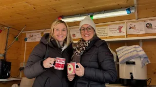 Zwei Frauen stehen draußen und lächeln, während sie festliche Tassen halten. Sie tragen Winterjacken und Schals, was auf eine kalte Umgebung hindeutet. Hinter ihnen befindet sich ein Regal mit einer Kaffeemaschine und einem Schild mit Preisen.