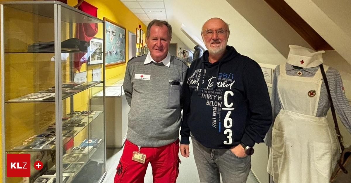 Zwei Männer stehen in einem Korridor, einer trägt ein graues Hemd und rote Hosen, der andere eine schwarze Kapuzenjacke. Sie stehen neben einer Glasvitrine mit Gegenständen und einer gelben Wand mit gerahmten Bildern.