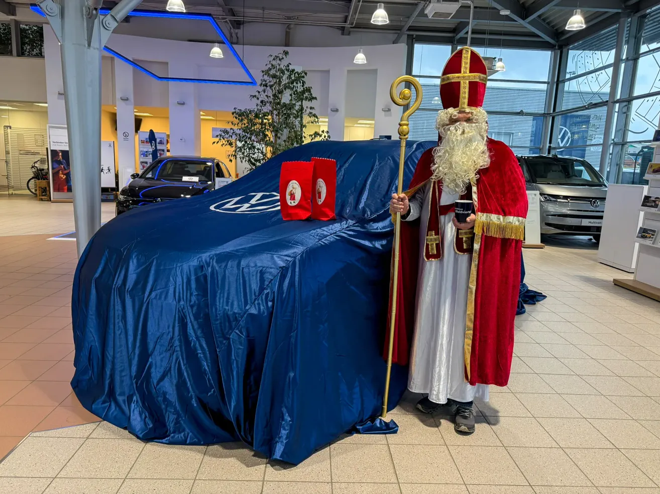 Ein Mann in einer Nikolaus-Verkleidung steht neben einem abgedeckten Auto, hält einen Stab und einen Becher in einer Ausstellungshalle.