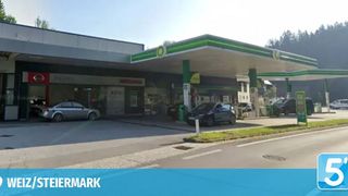 Eine BP-Tankstelle in Weiz, Steiermark, mit mehreren geparkten Autos, eines davon ist ein silbernes Auto mit zerbrochener Scheibe. Die Tankstelle hat grüne und weiße Schilder und befindet sich an einer Straße mit einem grasbewachsenen Mittelstreifen.