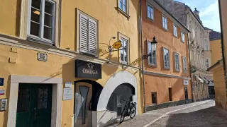 Eine schmale Kopfsteinpflasterstraße mit einem Fahrrad vor dem Omega Club & Bar. Das Gebäude hat gelbe Wände, weiße Fensterläden und ein schwarzes Vordach mit Omega-Marke.