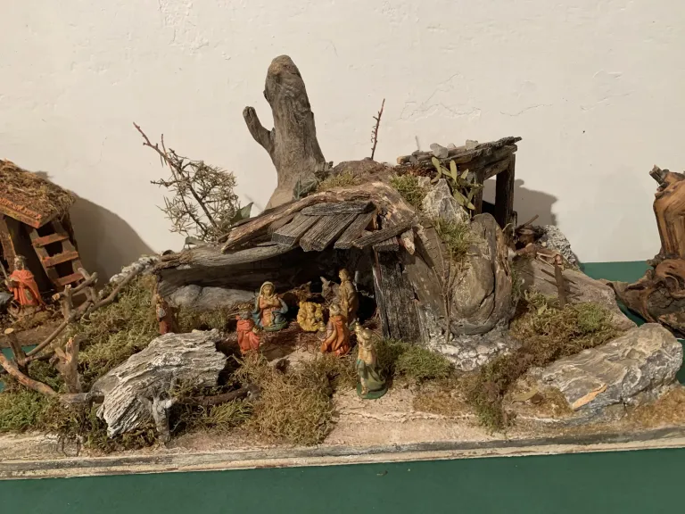 Eine Miniatur-Krippenszene mit einem Krippenstall mit Figuren in einer rustikalen Holzhütte, umgeben von Steinen, Moos und Zweigen auf einer grünen Oberfläche.