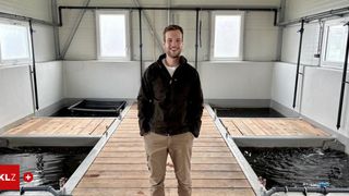 Ein Mann steht auf einem hölzernen Steg in der Nähe von Fischbecken und lächelt für ein Foto. Der Steg hat Geländer, und es gibt drei mit Wasser gefüllte Becken. Der Bereich hat weiße Wände und Fenster.