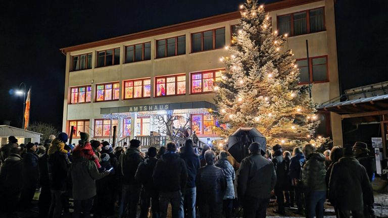 In der Nacht versammelt sich eine Menschenmenge vor einem Gebäude, 'Amtshaus', mit beleuchteten Fenstern. Ein hoher, beleuchteter Weihnachtsbaum steht in der Nähe.