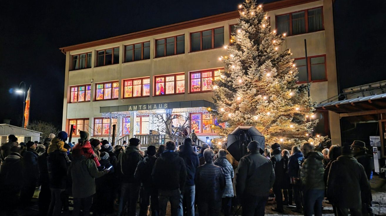 In der Nacht versammelt sich eine Menschenmenge vor einem Gebäude, 'Amtshaus', mit beleuchteten Fenstern. Ein hoher, beleuchteter Weihnachtsbaum steht in der Nähe.
