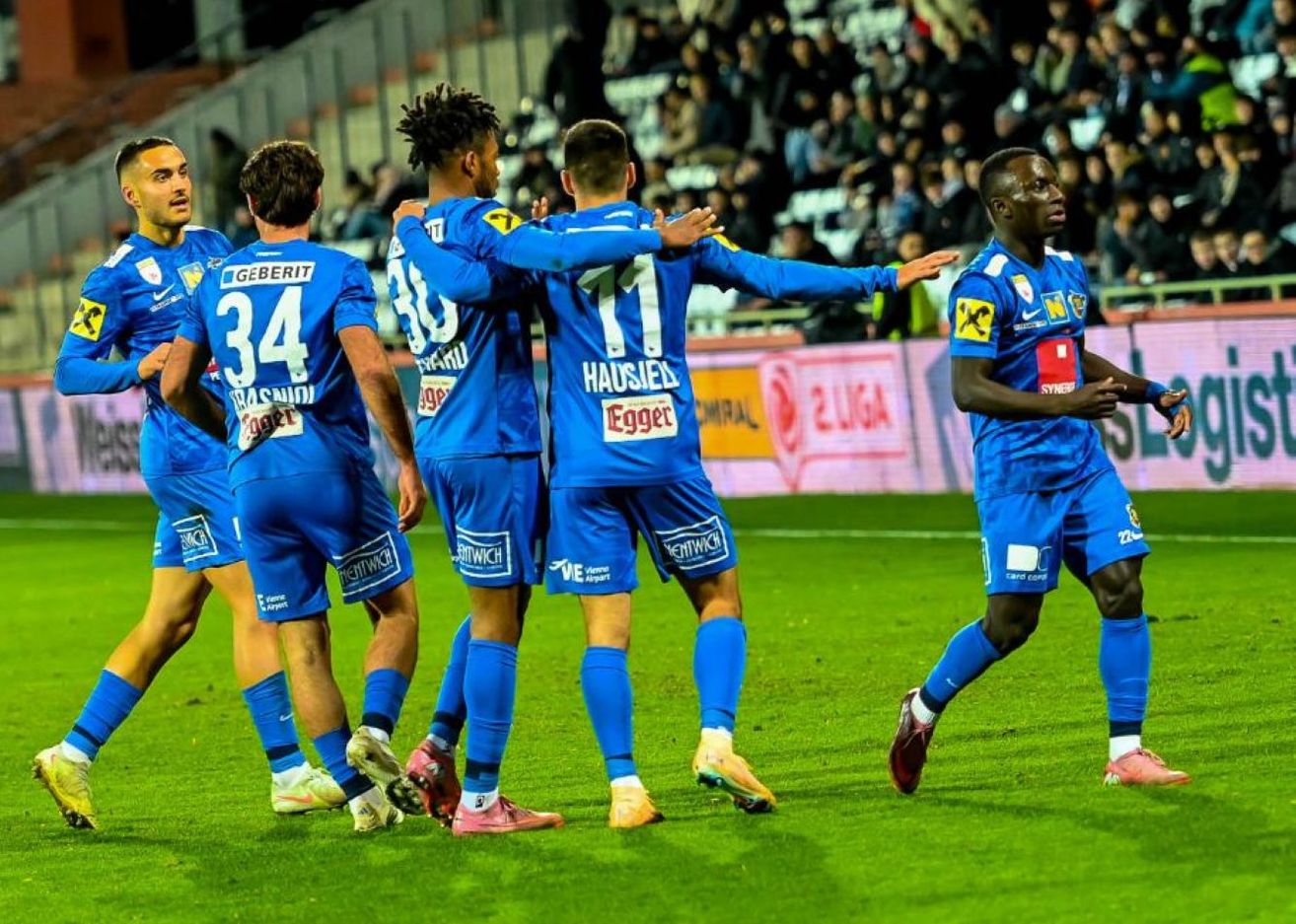A soccer team celebrates on the field, with players in blue uniforms, numbers 34, 30, and 11 visible. They stand close, showing camaraderie. The background includes a blurred audience and advertisement boards.