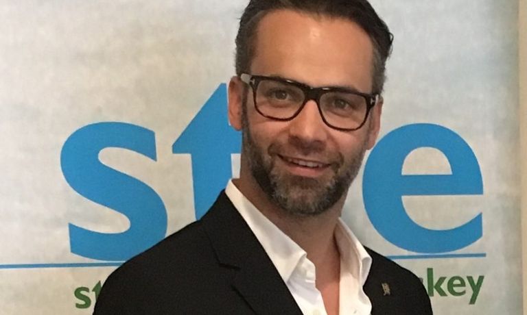 Ein Mann mit Brille, schwarzem Anzug und weißem Hemd steht lächelnd vor einem blau-weißen Banner.