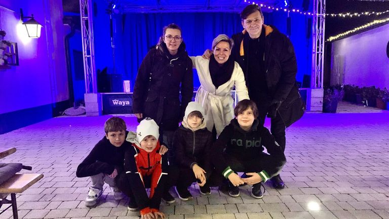 Eine Familie mit fünf Personen, darunter zwei Frauen und drei Jungen, posiert für ein Foto auf einem gefliesten Boden mit blauem Hintergrund. Die Frauen tragen Mäntel, eine davon einen Schal. Die Jungen tragen Hoodies und Turnschuhe. Ein blaues Licht beleuchtet die Szene.