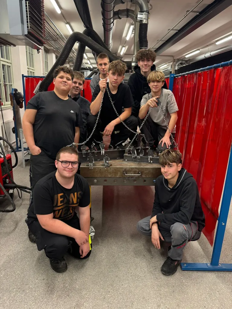 Eine Gruppe junger Männer posiert in einer Werkstatt, hält Ketten und lächelt in die Kamera. Sie stehen um ein großes Metallobjekt herum.