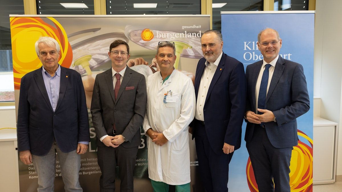 Fünf Männer in Anzügen stehen vor einem Krankenhausbanner. Die zentrale Figur trägt einen weißen Laborkittel, während andere Krawatten und Brillen tragen.