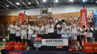 Die Basketballmannschaft feiert den Gewinn des Pokals mit der Aufschrift Üsterreichischer Meister 2025. Sie tragen Medaillen, weiße T-Shirts und Sneakers. Sie stehen vor einem blauen Banner mit der Werbung von Win2day und Superliga.