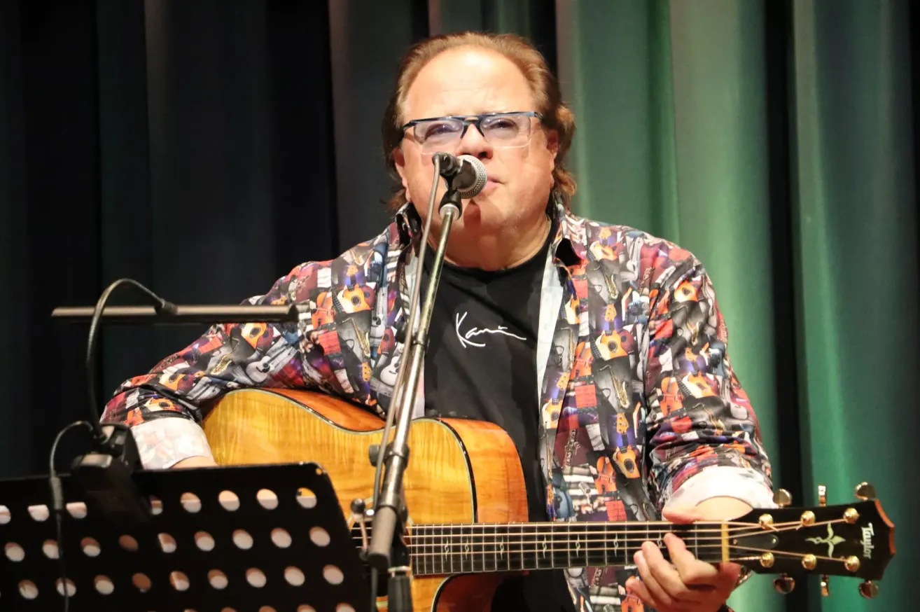 Ein Mann mit Brille spielt eine Gitarre und singt in ein Mikrofon auf der Bühne. Der Mann trägt ein farbenfrohes Hemd mit Drucken von Musikinstrumenten.