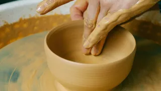 Die Hände einer Person formen eine Tonschale auf einer Töpferscheibe. Die Hände sind mit Tonstauß bedeckt. Die Schale steht auf einer Drehplatte, und die Person konzentriert sich auf ihre Formgebung.