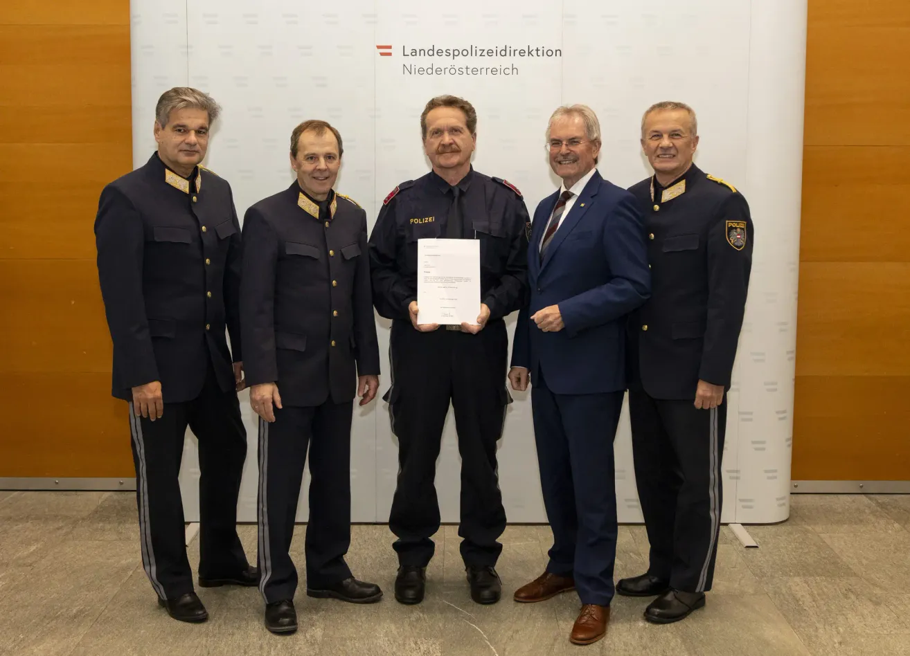 Fünf Männer in Uniformen stehen hinter einem weißen Hintergrund. Der zentrale Mann hält ein Dokument, und die anderen schauen ihn an. Der Hintergrund hat die Worte 'Landespolizeidirektion Niederosterreich'.