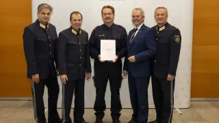 Fünf Männer in Uniformen stehen hinter einem weißen Hintergrund. Der zentrale Mann hält ein Dokument, und die anderen schauen ihn an. Der Hintergrund hat die Worte 'Landespolizeidirektion Niederosterreich'.