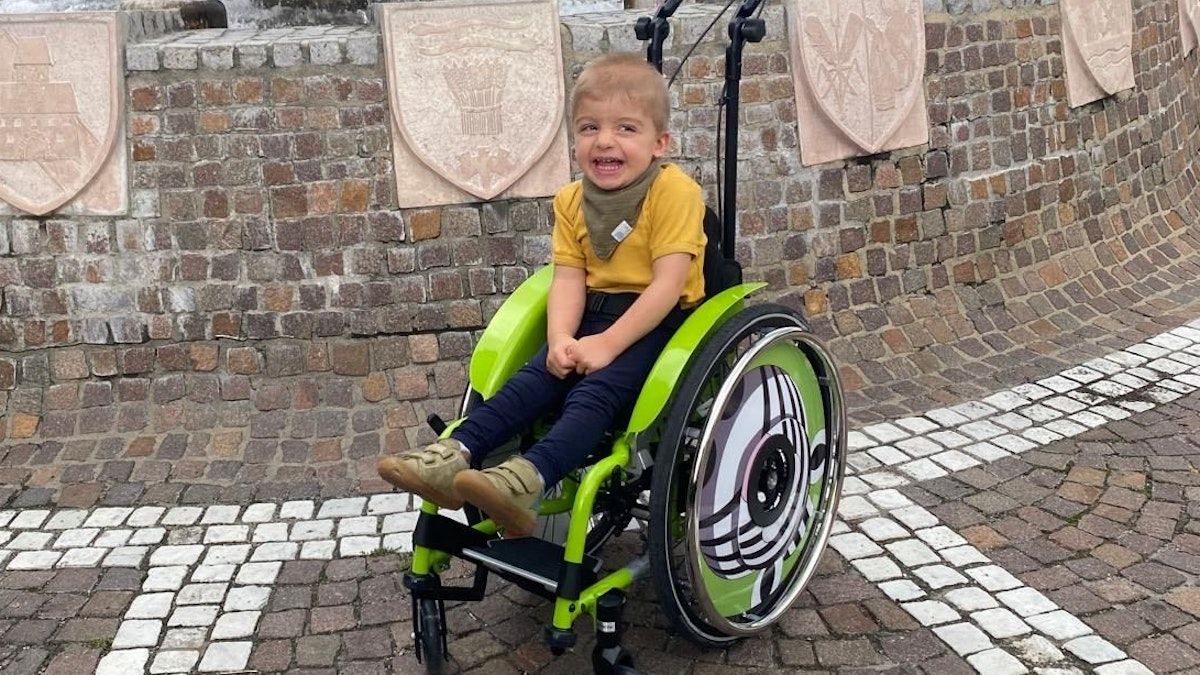 Ein Junge in einem gelben T-Shirt und blauen Hosen sitzt in einem grünen Rollstuhl mit einer Ziegelmauer und Schilden im Hintergrund.