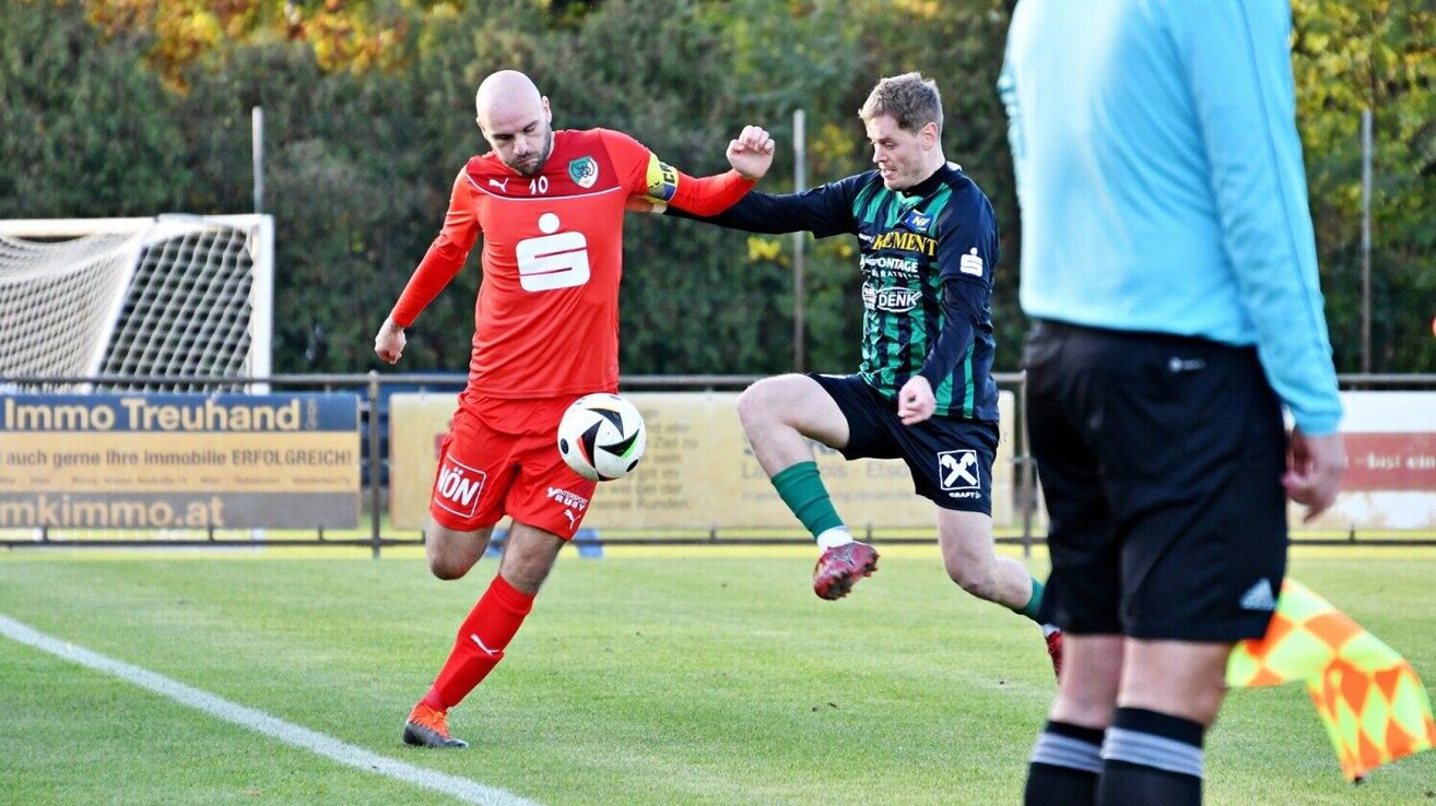 Zwei Fußballspieler konkurrieren auf einem Feld; einer in Rot mit Nummer 10, der andere in Grün und Blau. Der Spieler in Rot tritt den Ball, während der Spieler in Grün und Blau verteidigt.