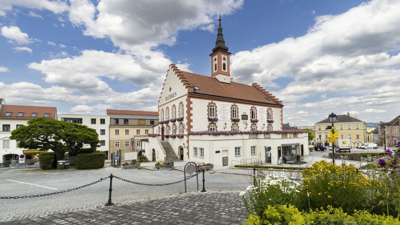 Ein historisches Gebäude mit Turm und Spitzturm steht auf einem Stadtplatz. Das Gebäude hat ein braunes Dach und weiße Wände mit Fenstern, die mit lila Blumen geschmückt sind.