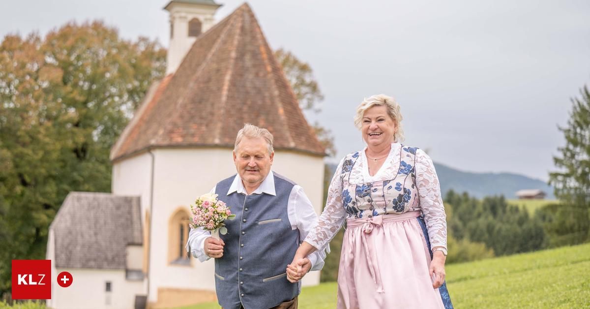 Ein älteres Paar, lächelnd, hält sich an den Händen, während es auf einem Feld spazieren geht, mit einer Kirche im Hintergrund. Der Mann hält einen Blumenstrauß.