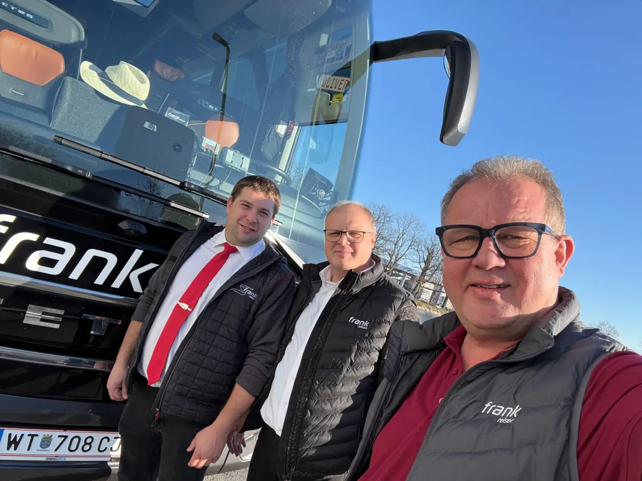 Drei Männer stehen vor einem Bus, zwei in schwarzen Jacken, einer in einem roten Hemd. Der Mann auf der linken Seite trägt eine rote Krawatte, während der in der Mitte eine Brille trägt. Hinter ihnen steht ein Bus mit dem Wort 'OLIVER' auf der Seite.
