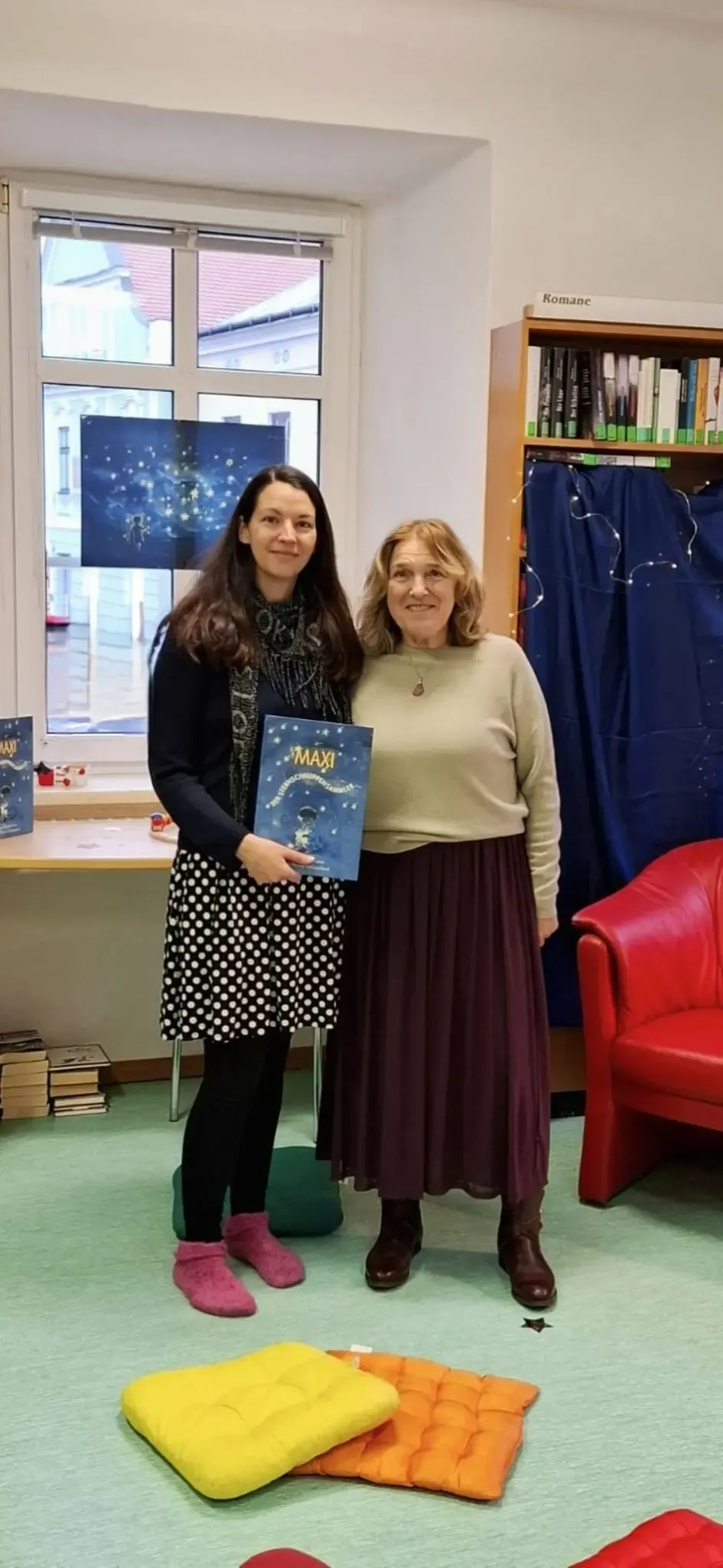 Zwei Frauen stehen in einem Raum, eine hält ein Buch mit dem Titel 'Maxi', sie lächeln für ein Foto mit einem Schreibtisch, einem Stuhl und Büchern im Hintergrund.