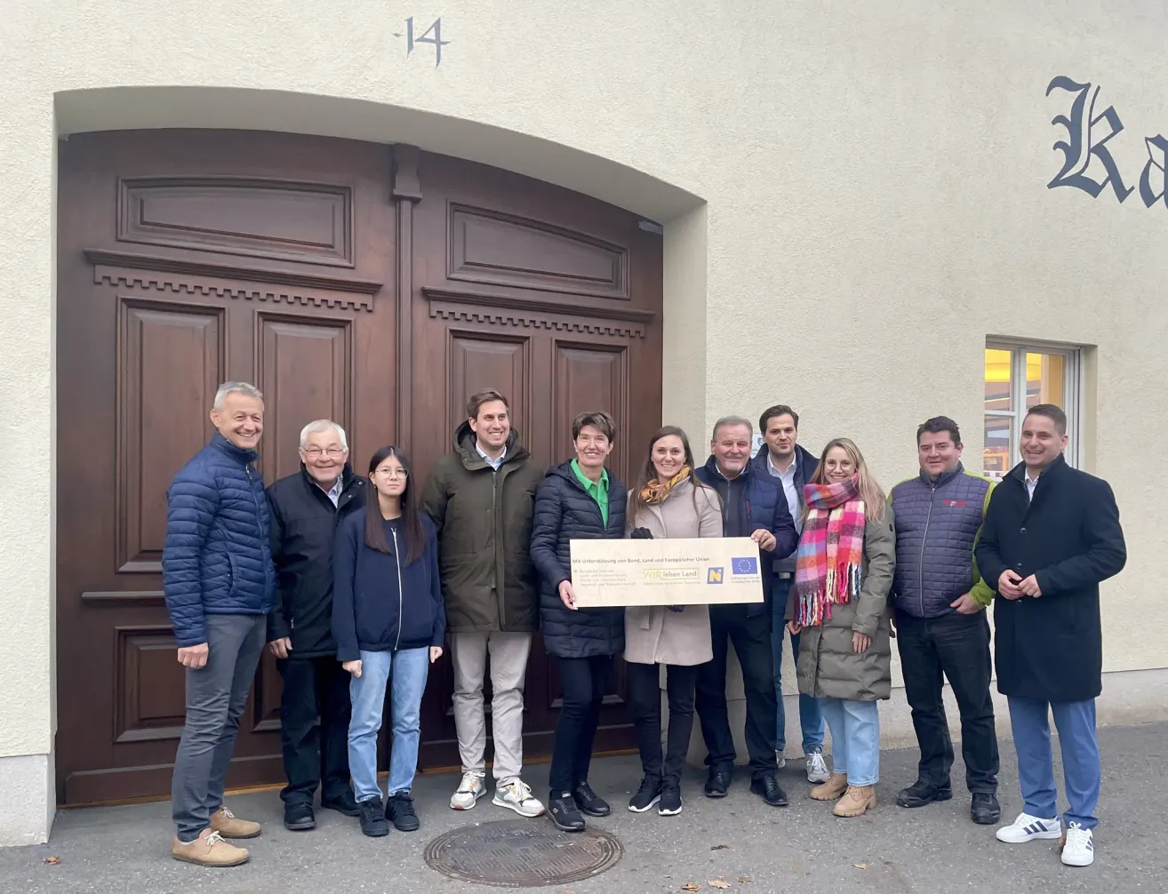Eine Gruppe von Menschen steht vor einem Gebäude und posiert für ein Foto. Sie halten einen großen Scheck. Der Mann in der Mitte trägt ein grünes Shirt. Über ihnen steht die Nummer 14.