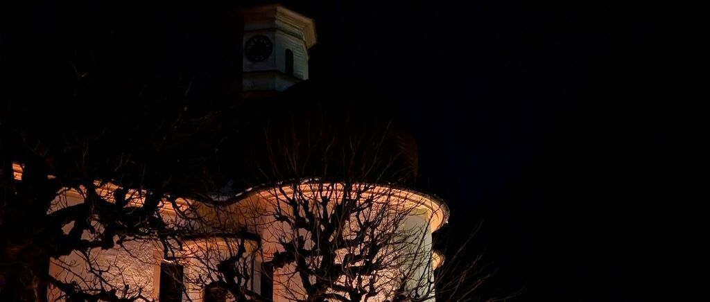 Eine Kirche mit einem Glockenturm ist bei Nacht beleuchtet. Die Architektur des Gebäudes umfasst eine Kuppel und einen Turm. Das Licht hebt die Kuppel und den Turm hervor.