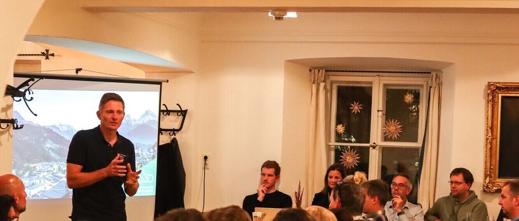 Ein Mann hält eine Präsentation in einem Raum mit einer Projektionsleinwand hinter ihm. Er steht vor einem sitzenden Publikum.