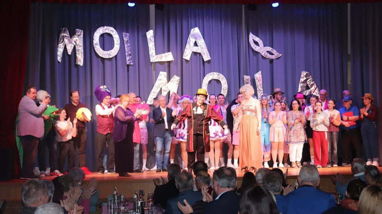 Eine Gruppe von Menschen auf der Bühne führt ein Theaterstück auf, das Publikum schaut zu. Ein Banner hinter ihnen trägt den Schriftzug 'OLI LA MOLI'. Einige der Darsteller tragen Kostüme und Hüte.