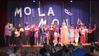 Eine Gruppe von Menschen auf der Bühne führt ein Theaterstück auf, das Publikum schaut zu. Ein Banner hinter ihnen trägt den Schriftzug 'OLI LA MOLI'. Einige der Darsteller tragen Kostüme und Hüte.