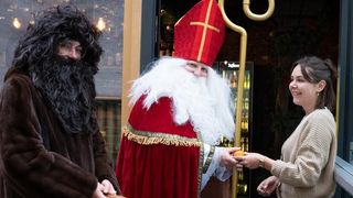 Ein Mann in einem Weihnachtsmannkostüm gibt einer Frau eine Orange. Ein anderer Mann in einem Pelzmantel steht neben ihm. Sie stehen vor einem Geschäft.