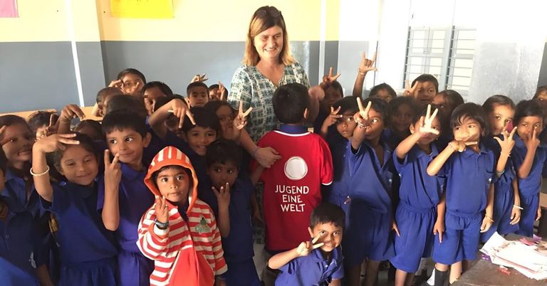 Eine lächelnde Frau steht inmitten einer Gruppe von Kindern, die alle Friedenszeichen machen. Die Kinder tragen blaue Uniformen und die Frau trägt ein grünes Oberteil. Das Kind in der Mitte trägt ein rotes Hemd mit weißem Text.