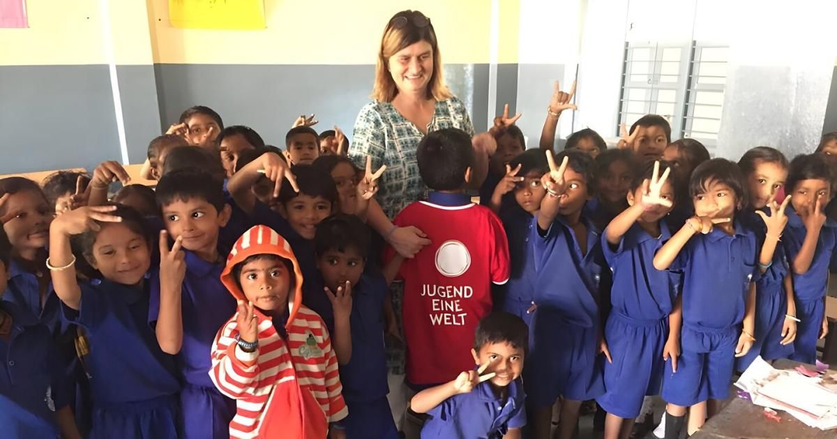 Eine lächelnde Frau steht inmitten einer Gruppe von Kindern, die alle Friedenszeichen machen. Die Kinder tragen blaue Uniformen und die Frau trägt ein grünes Oberteil. Das Kind in der Mitte trägt ein rotes Hemd mit weißem Text.