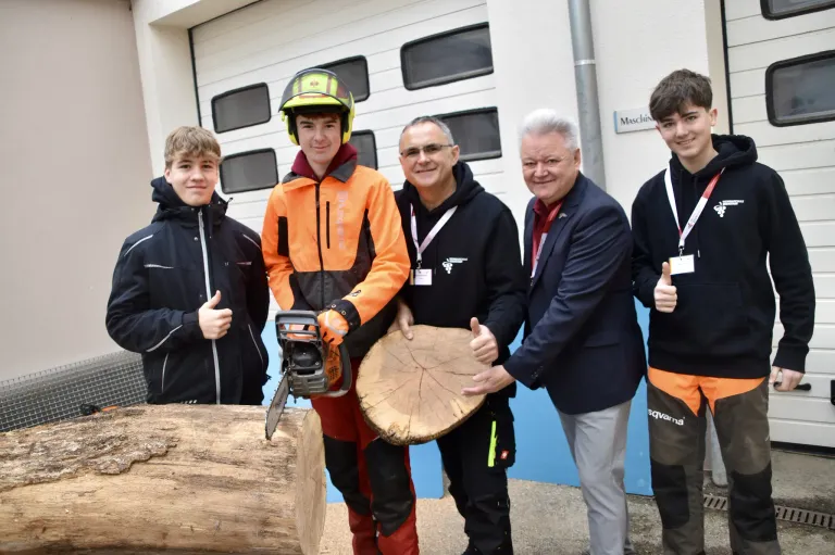 Fünf Männer stehen vor einem Gebäude mit einem Baumstamm im Vordergrund. Ein Mann hält eine Kettensäge, während andere Daumen hochhalten.