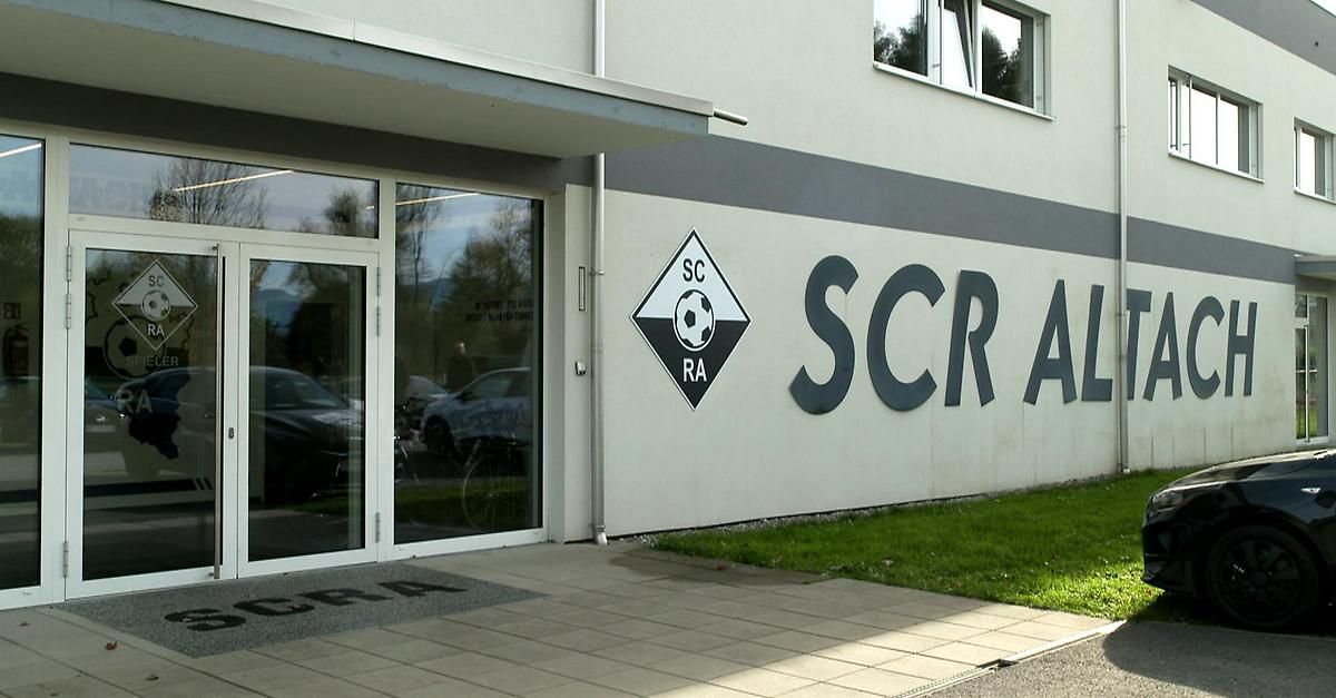 Der Eingang eines Gebäudes mit einer Glastür und einem Schild, das 'SCR A' steht. Ein Logo mit 'SC RA' ist an der Wand. Fahrräder sind draußen geparkt.