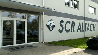 Der Eingang eines Gebäudes mit einer Glastür und einem Schild, das 'SCR A' steht. Ein Logo mit 'SC RA' ist an der Wand. Fahrräder sind draußen geparkt.