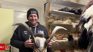 Ein Mann in einem schwarzen Hoodie mit Hörnern steht in einem Raum mit Regalen voller Tiermasken und Hörnern. Er lächelt und hält eine maskierte Hornmaske.