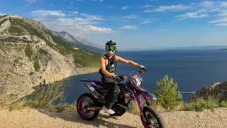 Eine Person auf einem violetten Motorrad mit Berg- und Ozeanhintergrund. Der Fahrer trägt Helm, Schutzbrille und Handschuhe.