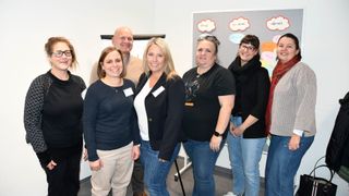 Eine Gruppe von Frauen und einem Mann posiert für ein Bild in einem Raum mit grauem Hintergrund und Haftnotizen.