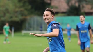Ein Fußballspieler in einem blauen Trikot lächelt und gestikuliert auf einem Feld, mit einem verschwommenen Hintergrund von Zaun und Bäumen.