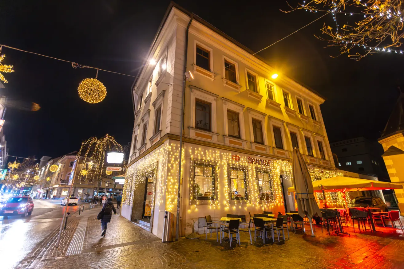 Ein Gebäude mit einem Restaurant im Erdgeschoss ist nachts hell beleuchtet. Der Eingang ist mit Lichtern dekoriert, und draußen sind Tische und Stühle aufgestellt. Eine Person mit Rucksack läuft auf dem Bürgersteig.