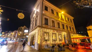 Ein Gebäude mit einem Restaurant im Erdgeschoss ist nachts hell beleuchtet. Der Eingang ist mit Lichtern dekoriert, und draußen sind Tische und Stühle aufgestellt. Eine Person mit Rucksack läuft auf dem Bürgersteig.