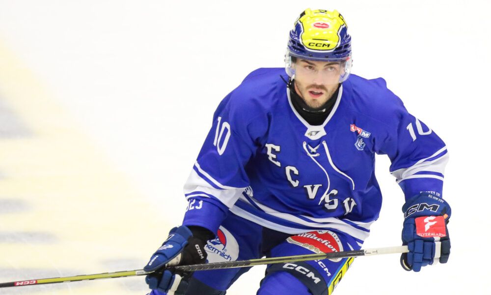 Ein Eishockeyspieler in einem blauen Trikot mit der Nummer 10 auf dem Rücken fährt auf dem Eis. Er hält einen Schläger in der rechten Hand.