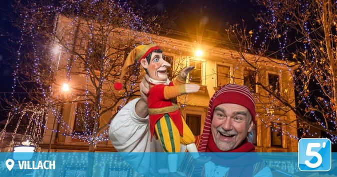 Ein Mann hält eine bunte Puppe vor einem Gebäude mit festlichen Lichtern und lächelt in die Kamera. Bäume mit Lichtern und ein Logo sind im Hintergrund zu sehen.