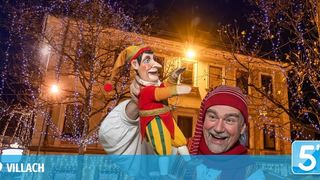 Ein Mann hält eine bunte Puppe vor einem Gebäude mit festlichen Lichtern und lächelt in die Kamera. Bäume mit Lichtern und ein Logo sind im Hintergrund zu sehen.