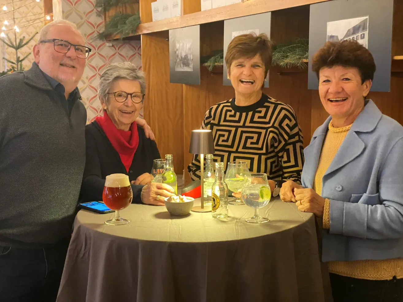 Vier Menschen sitzen um einen Tisch herum und lächeln. Zwei Frauen und ein Mann, wobei eine Frau ein Bierglas hält. Flaschen und Gläser stehen auf dem Tisch. Dahinter eine Holzwand mit gerahmten Bildern.